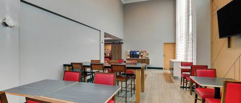 A modern dining area with multiple tables and red chairs, a flat-screen TV mounted on the wall, and a large window with curtains.