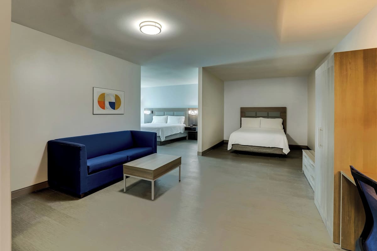 A modern hotel room with a blue sofa, a wooden coffee table, a bed with white linens, and a wardrobe.