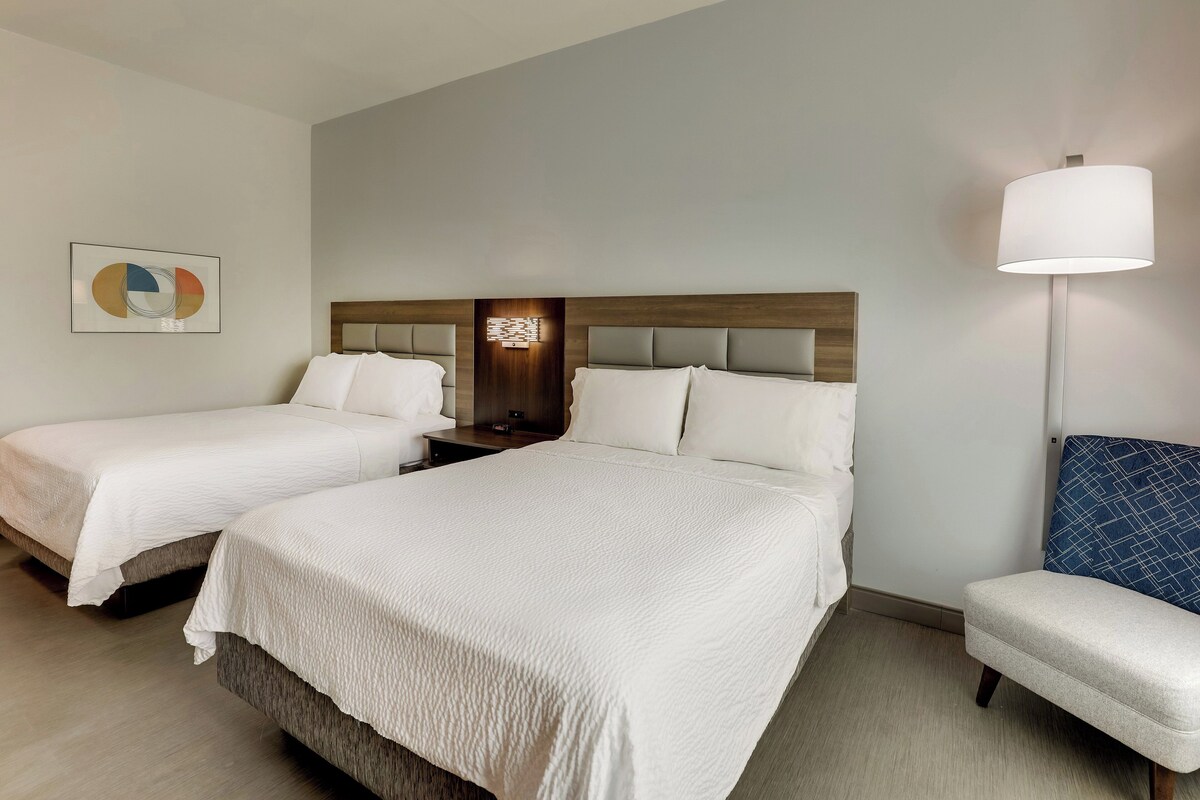 A hotel room with two beds, a nightstand, a lamp, and a chair.