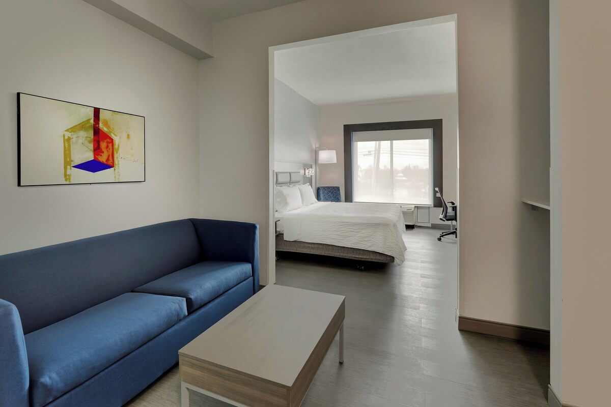 A modern hotel room with a blue sofa, a small wooden coffee table, a bed with white bedding, and a large window providing natural light.