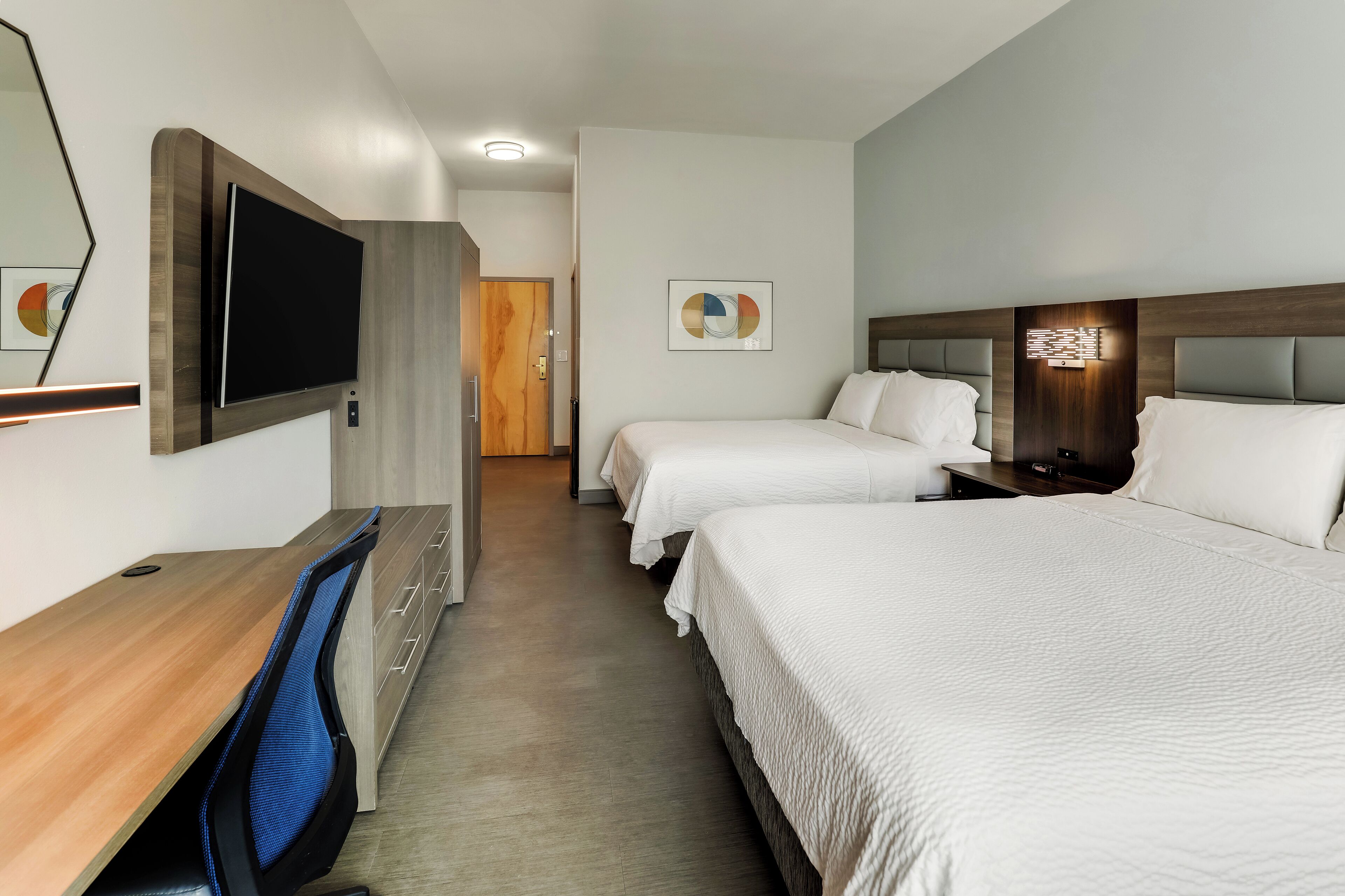 A hotel room with a desk, two beds, a TV, and a door leading to another room.