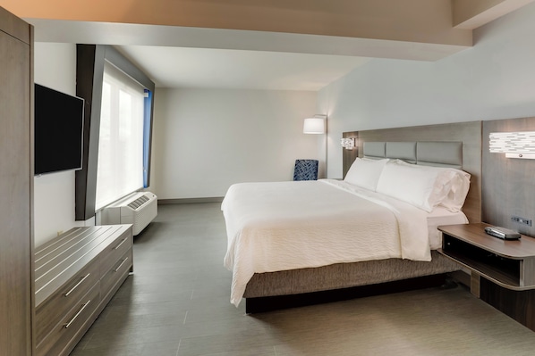 A modern hotel room with a large bed, a flat-screen TV mounted on the wall, a bedside table with a lamp, and a built-in heater.