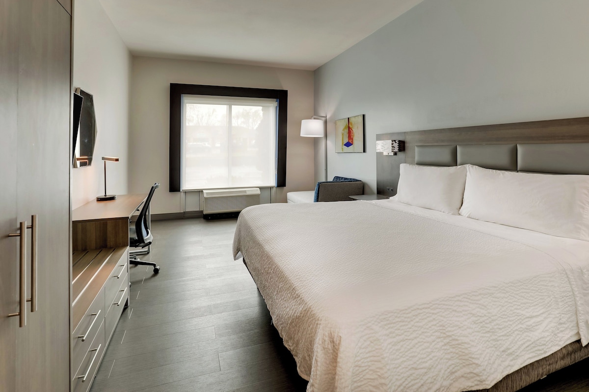 A hotel room with a large bed, a desk with a chair, a standing lamp, and a window.