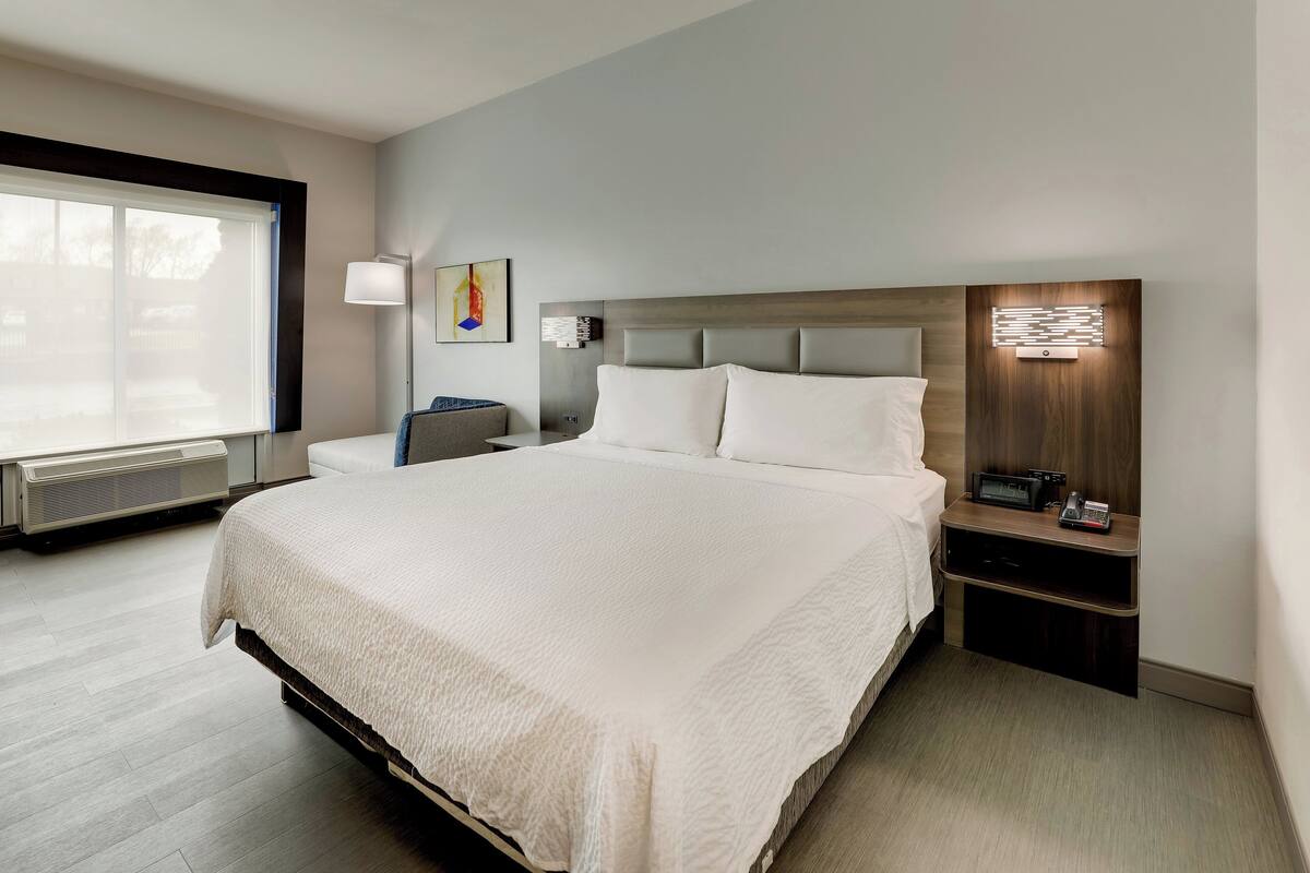 A modern hotel room with a large bed, a bedside table with a lamp, and a view of the outdoors through a large window.