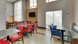 Un espace repas moderne, doté de plusieurs tables et chaises, d’un téléviseur à écran plat fixé au mur et de grandes fenêtres laissant entrer la lumière naturelle.