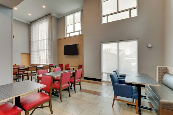 A modern dining area with multiple tables and chairs, a flat-screen TV mounted on the wall, and large windows providing natural light. - Holiday Inn Express & Suites Richmond by IHG (Richmond)