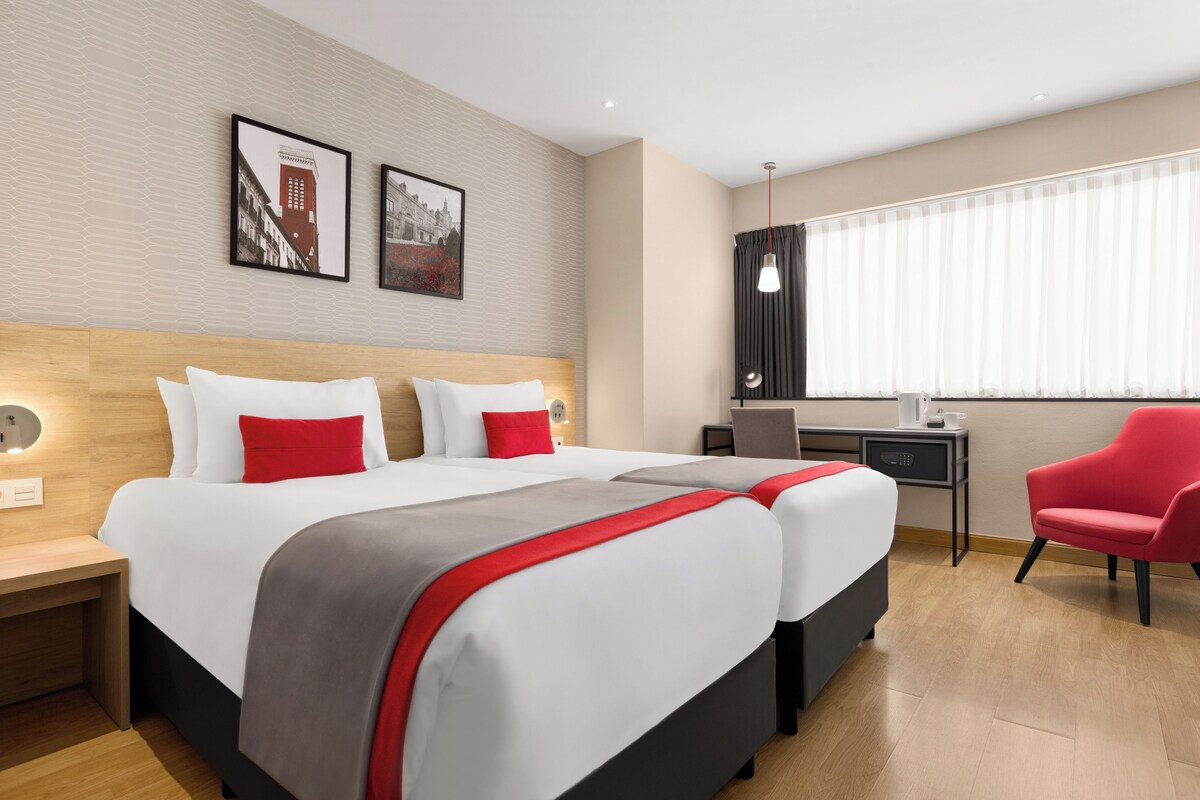 A hotel room with two beds, a desk, a chair, and a window with blinds.