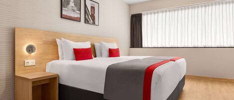 Una habitación de hotel moderna con cabecera de madera, mesita de noche con lámpara, cama grande con ropa blanca y detalles rojos, y ventana con cortinas de encaje.