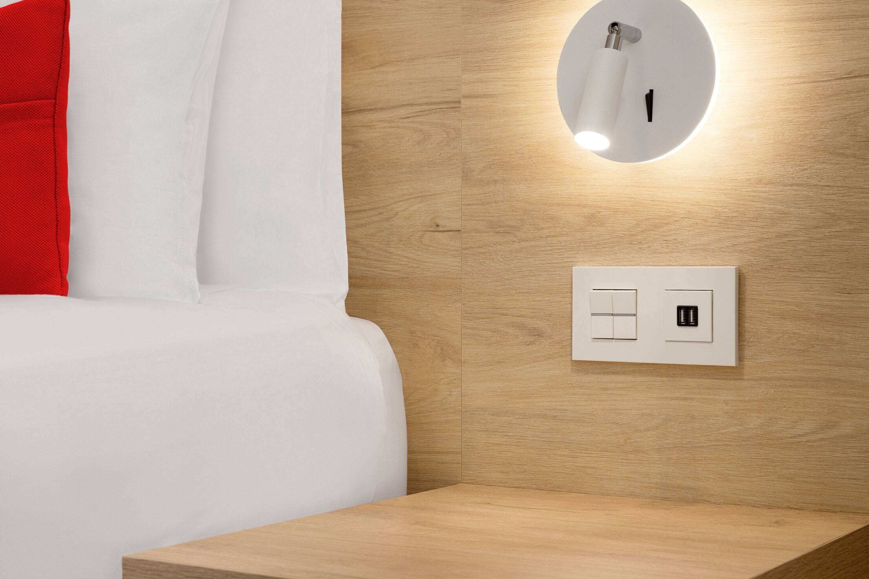 A hotel room with a wooden headboard, a bedside table, a white bed, a red pillow, a wall-mounted light fixture, and a power outlet.