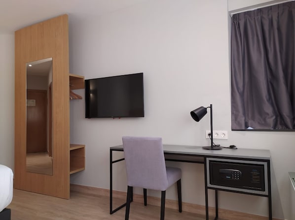 A modern room with a wooden wardrobe, a flat-screen TV mounted on the wall, a desk with a computer, a chair, and a window with curtains.