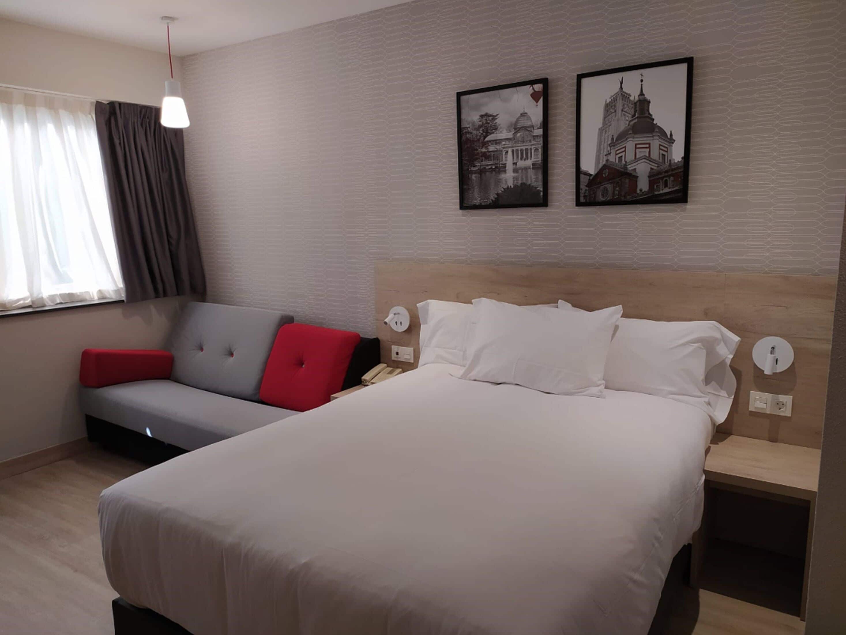 Una habitación de hotel moderna con una cama grande, un sofá gris con cojines rojos, una ventana con cortinas y dos cuadros en la pared.