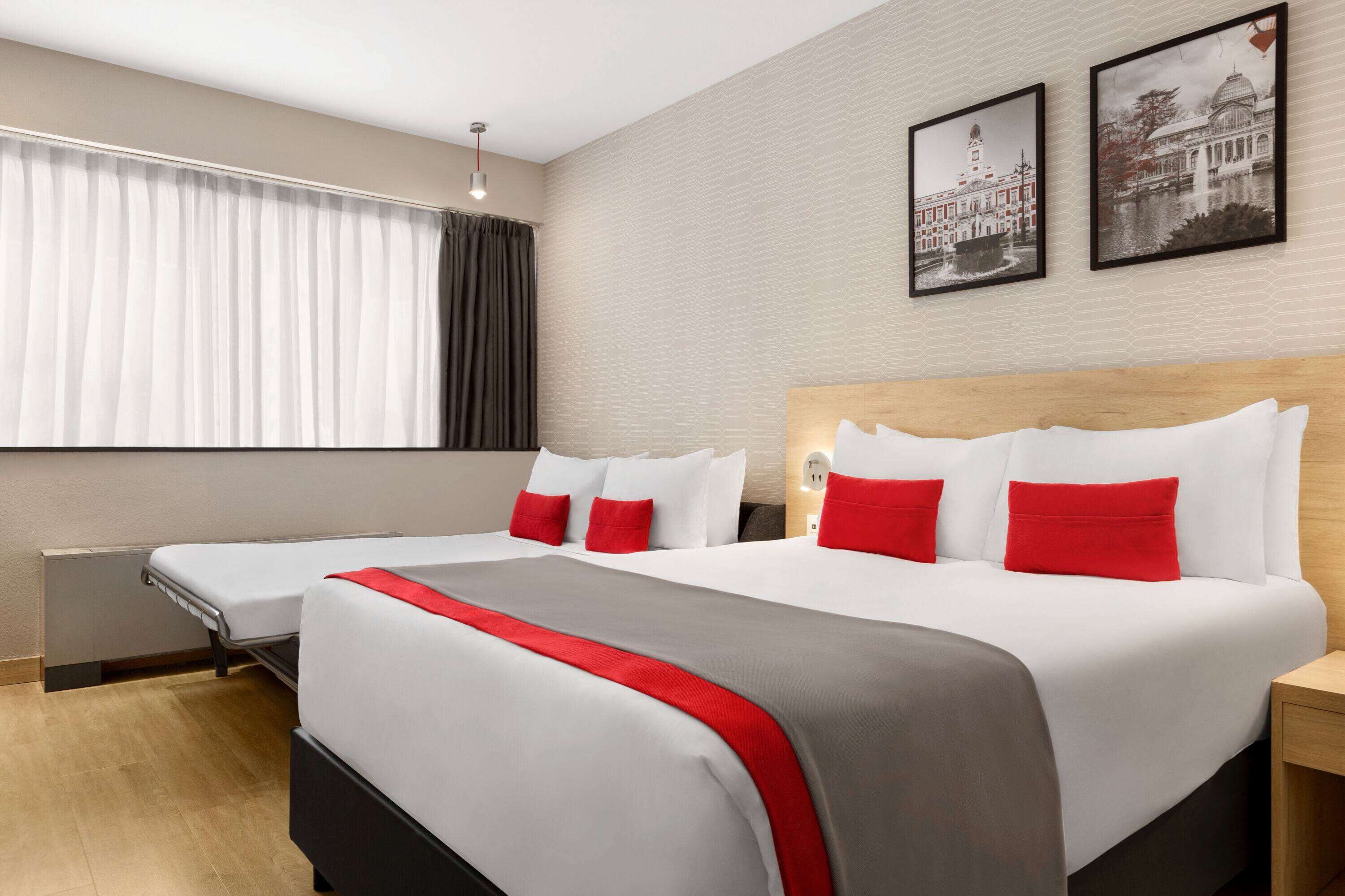 A hotel room with two beds, a wooden headboard, a red and grey bedspread, and two black-and-white framed pictures on the wall.