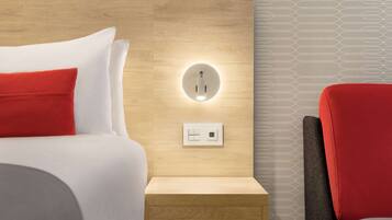 Una habitación de hotel moderna con cabecera de madera, una mesita de noche, una silla roja y un aplique de pared.