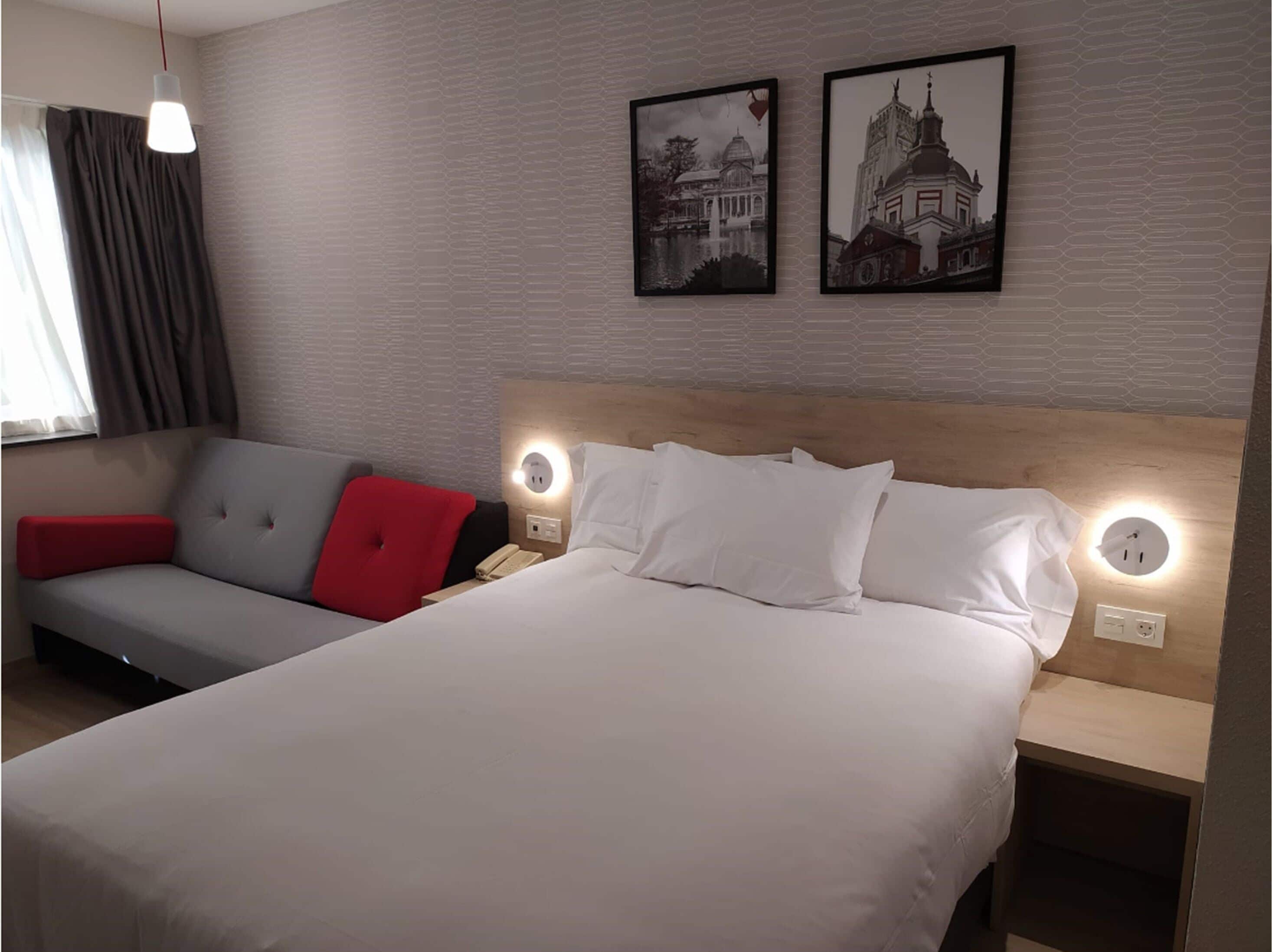 A hotel room with a bed, a grey sofa with a red pillow, a red chair, and two framed pictures on the wall.
