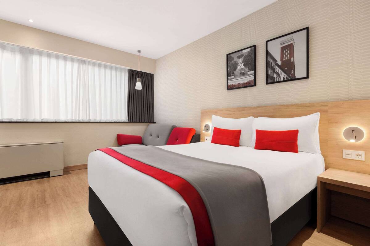 Ein modernes Hotelzimmer mit einem großen Bett, einem Nachttisch, einem Fenster mit Vorhängen und zwei gerahmten Bildern an der Wand.