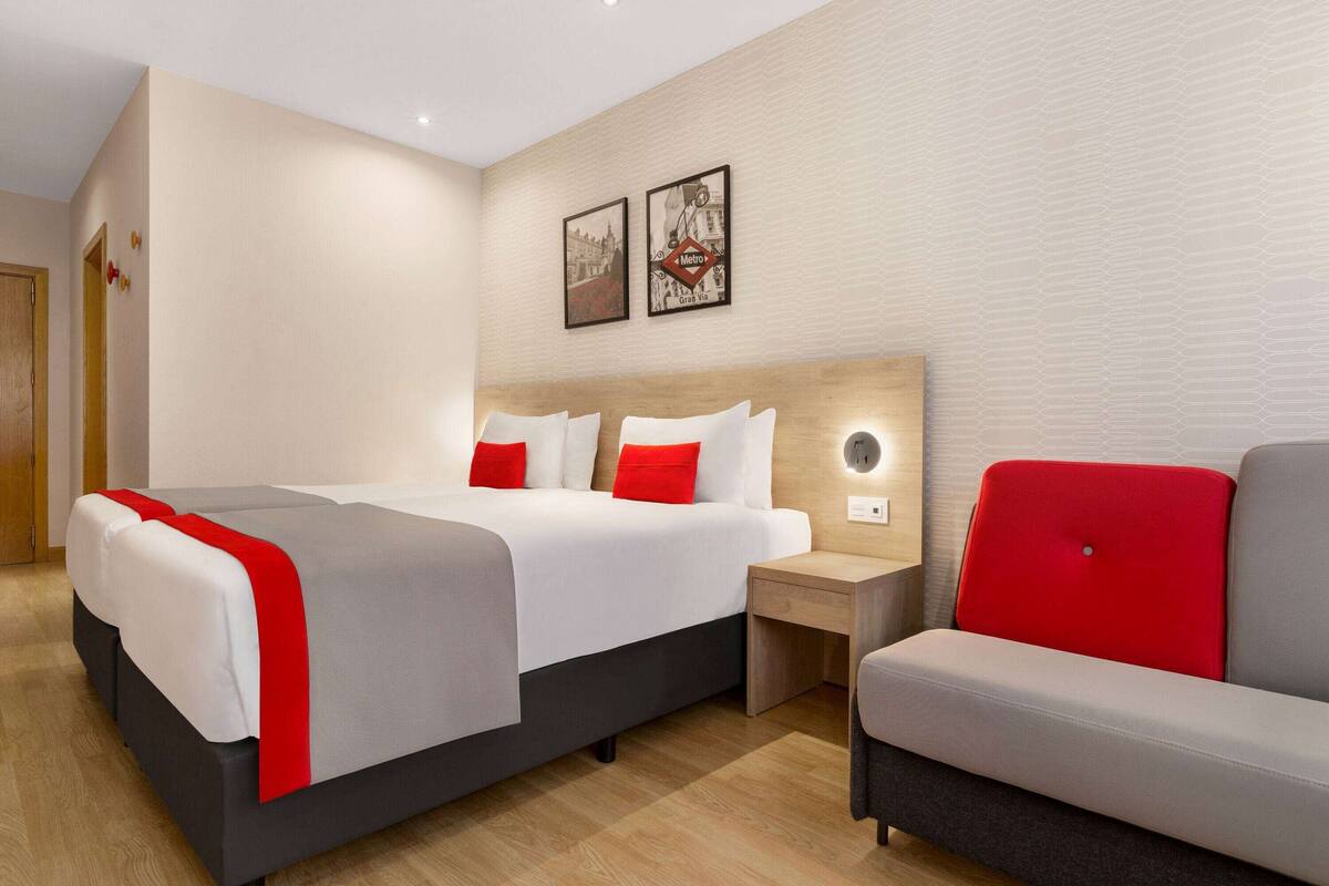 Ein modernes Hotelzimmer mit einem Bett, einem Nachttisch, einem rot-grauen Sofa und zwei gerahmten Bildern an der Wand.