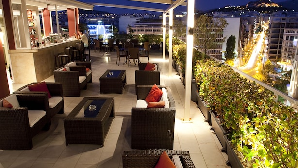 A rooftop terrace with wicker furniture, a bar area, and a view of the cityscape at night. - Crystal City Hotel (Athens)