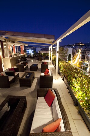 Una terraza en la azotea con mobiliario de mimbre, una zona de bar y vistas a la ciudad iluminada por la noche.