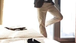 A person in a suit standing by a window with a tablet on the bed. - Crystal City Hotel (Athens)