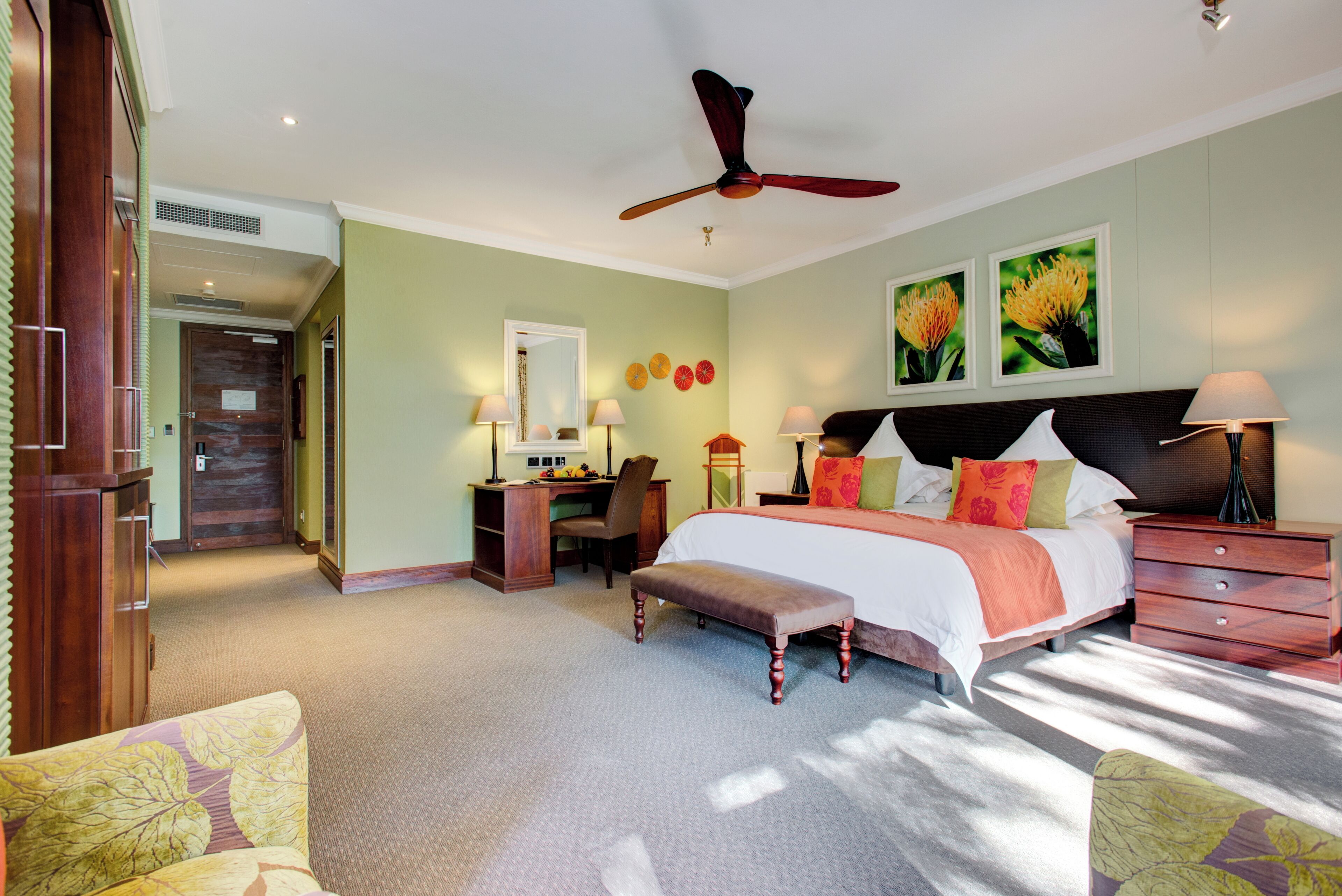 A bedroom with a bed, bedside tables, a desk, a chair, a ceiling fan, and artwork on the wall.