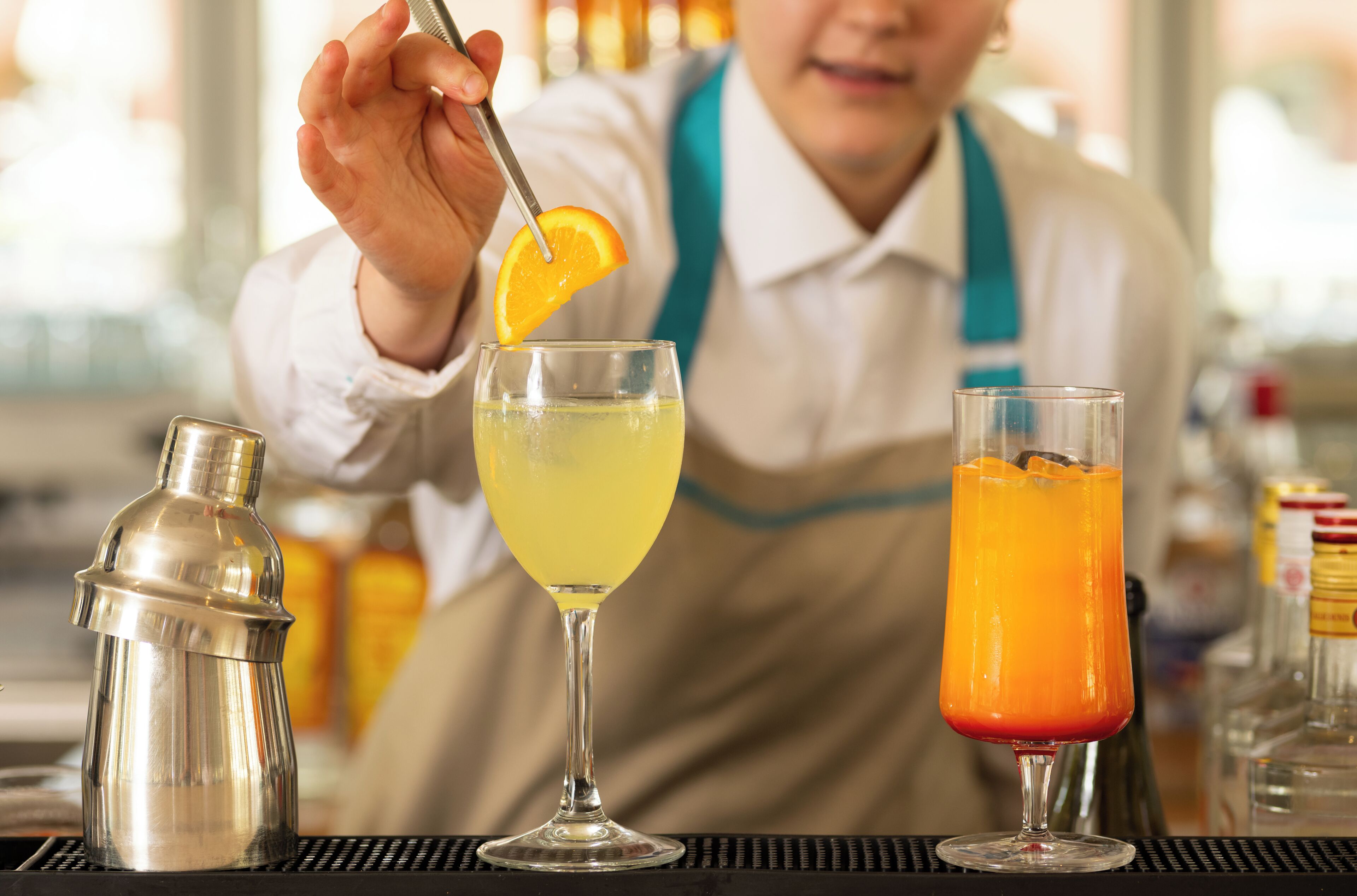 Um bartender preparando bebidas com um coqueteleira, um coquetel amarelo e uma bebida de laranja.
