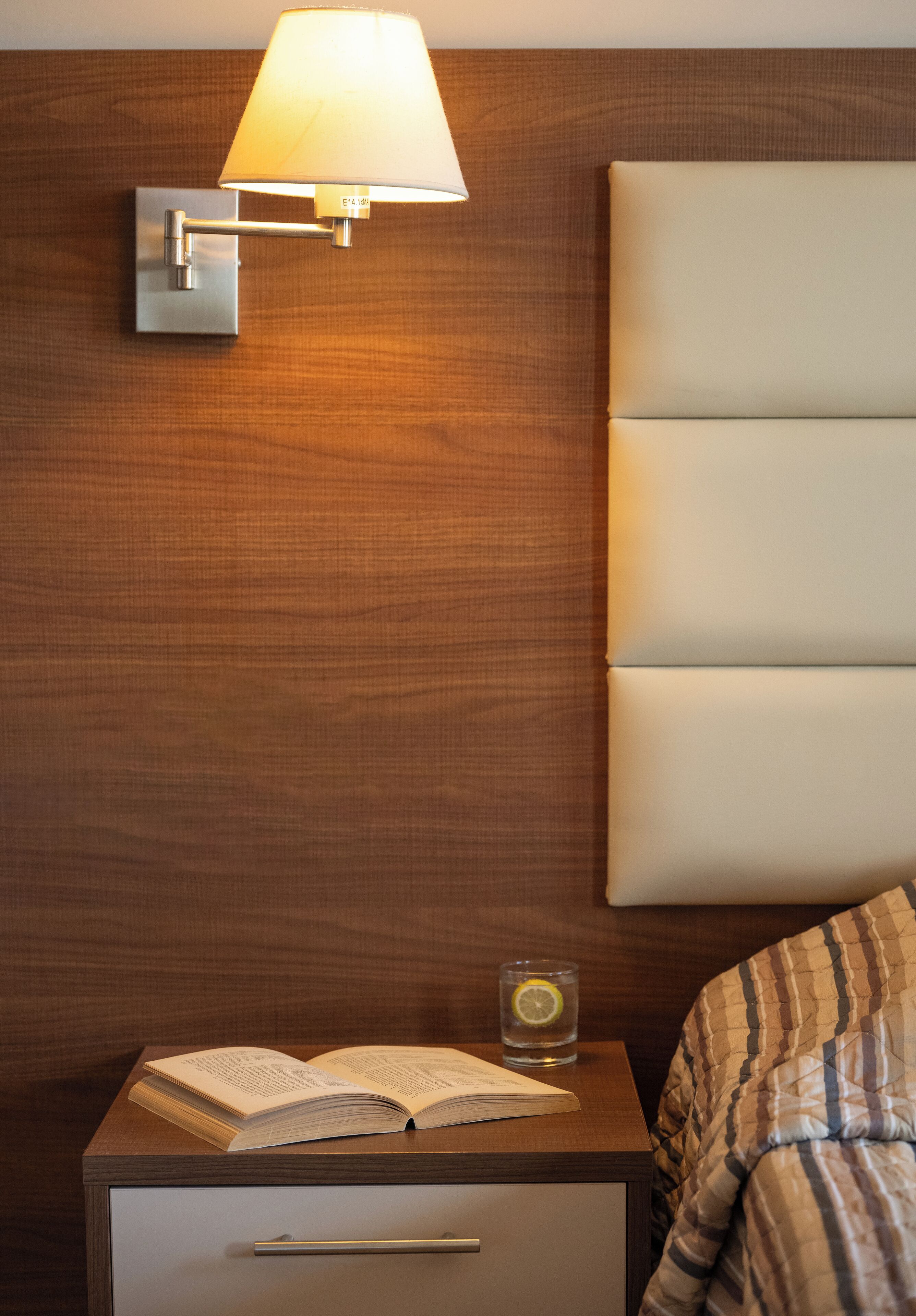 Un lit bien fait, recouvert d’une couverture à rayures, une table de chevet avec un livre ouvert et un verre d’eau, et une lampe fixée au mur.