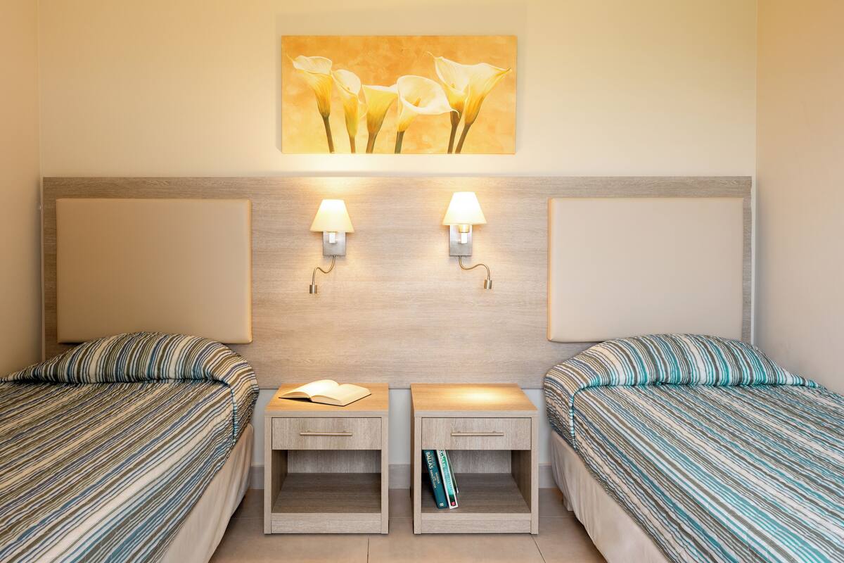 A hotel room with two beds, each with striped bedding, a nightstand with a book and a pen, and a wall-mounted lamp.