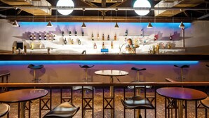 Una zona de bar con un cantinero preparando bebidas, una pared llena de botellas de licor y varias mesas y sillas redondas.