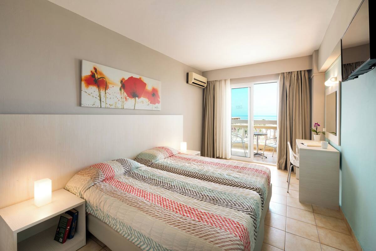 A hotel room with two beds, a desk, a chair, a window with curtains, and a painting on the wall.