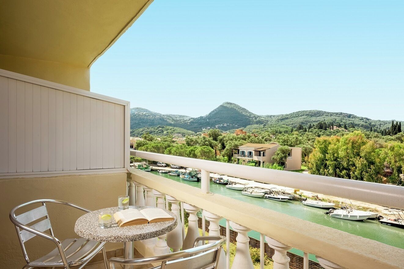 Un balcón con mesa y sillas, con vista a un río con barcos y un paisaje montañoso.