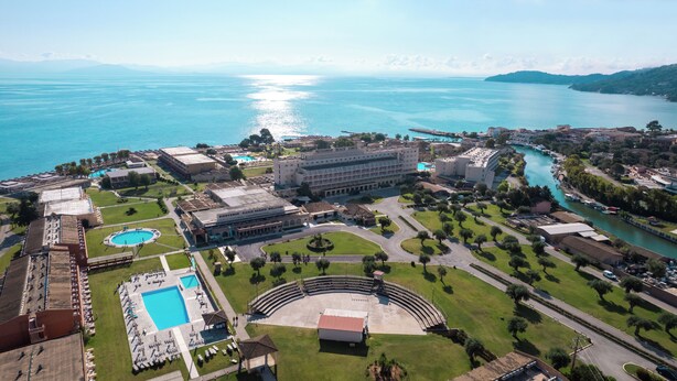 Vista aérea de un complejo turístico costero con un gran edificio identificado como “MESSONGHI BEACH CORFU”, una piscina y un río que discurre junto a la propiedad.