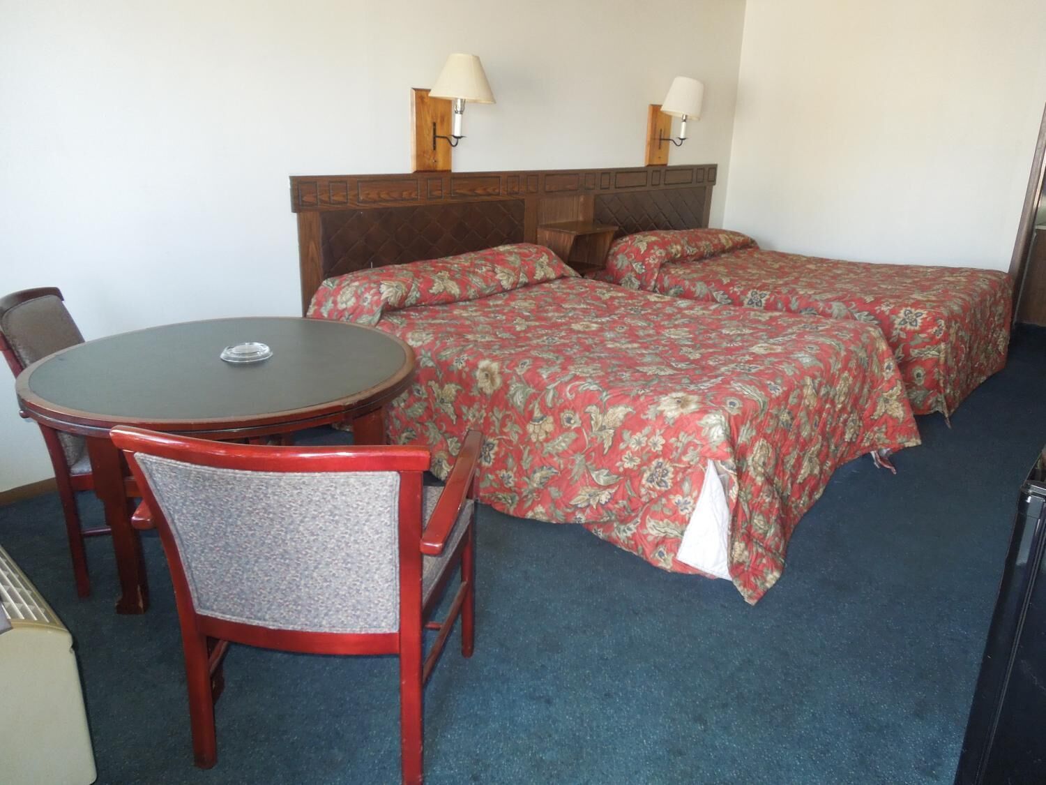 Habitación de hotel con cabecera de madera, dos camas individuales con ropa de cama floral, una mesa redonda con una silla y dos lámparas fijadas a la pared.