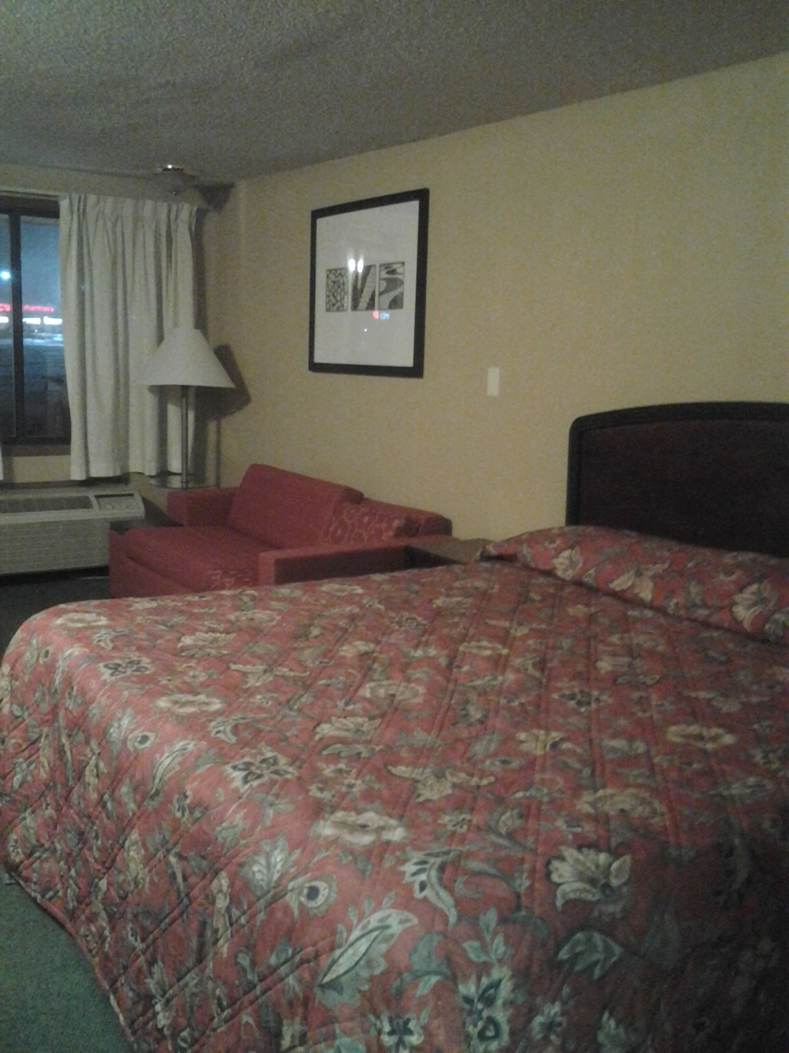 A hotel room with a large bed, a red chair, a framed picture on the wall, and a window with curtains.