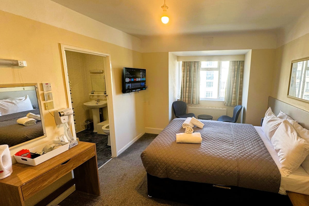 Ein Hotelzimmer mit Bett, Schreibtisch, Spiegel, Fernseher und einem Fenster mit Vorhängen sowie Blick auf Gebäude.