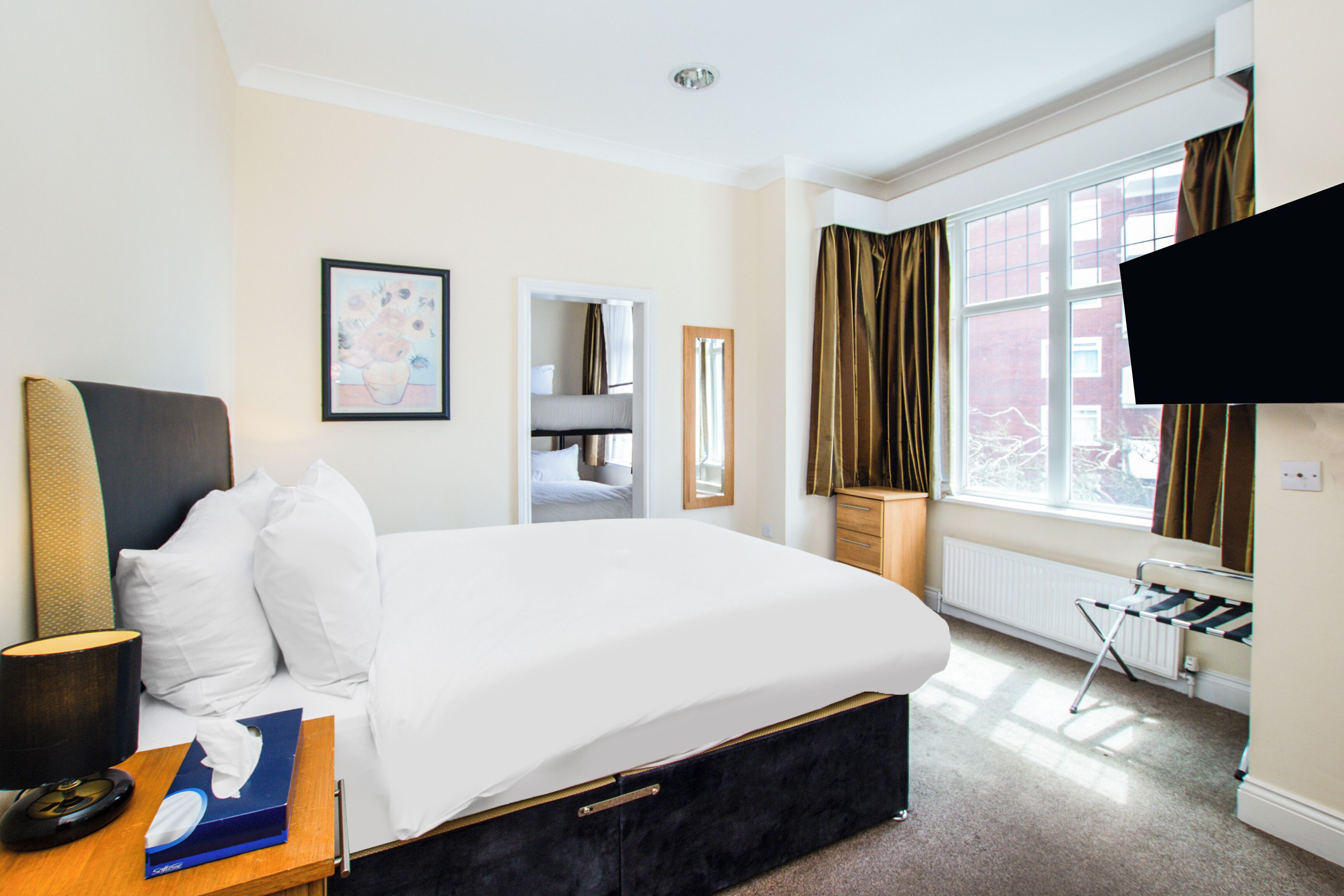 A hotel room with a bed, bedside table, lamp, and a view of the outside.