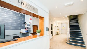 Ein Empfangsbereich mit Empfangstresen, einem Computer, einem Telefon und einer Pflanze. Eine Treppe führt zu einer anderen Etage.