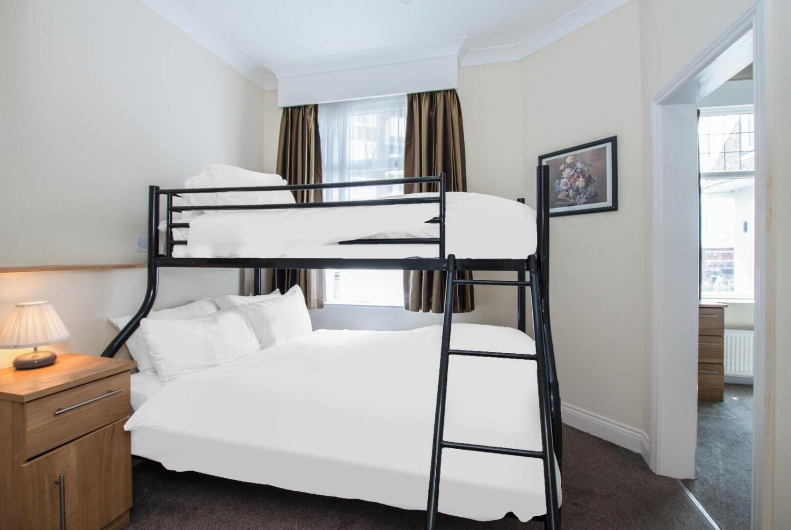 A bunk bed with a bedside table and lamp, a framed picture on the wall, and a view of the outside through a window.