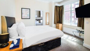 Ein Hotelzimmer mit Bett, Nachttisch, Lampe, Kunstwerk, Fenster mit Vorhängen, Heizkörper und Fernseher.