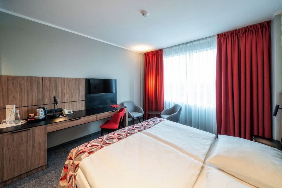 Une chambre d’hôtel avec un lit, un bureau, des chaises, une télévision et une fenêtre avec des rideaux.