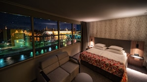 Une chambre d’hôtel dotée d’une grande fenêtre donnant sur la ville de nuit, d’un lit avec une couvre-lit à motifs géométriques, d’un canapé et d’une petite table avec une lampe.