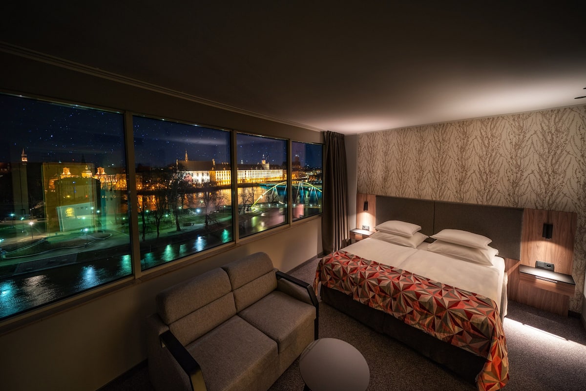 Une chambre d’hôtel dotée d’une grande fenêtre donnant sur la ville de nuit, d’un lit avec une couvre-lit à motifs géométriques, d’un canapé et d’une petite table avec une lampe.
