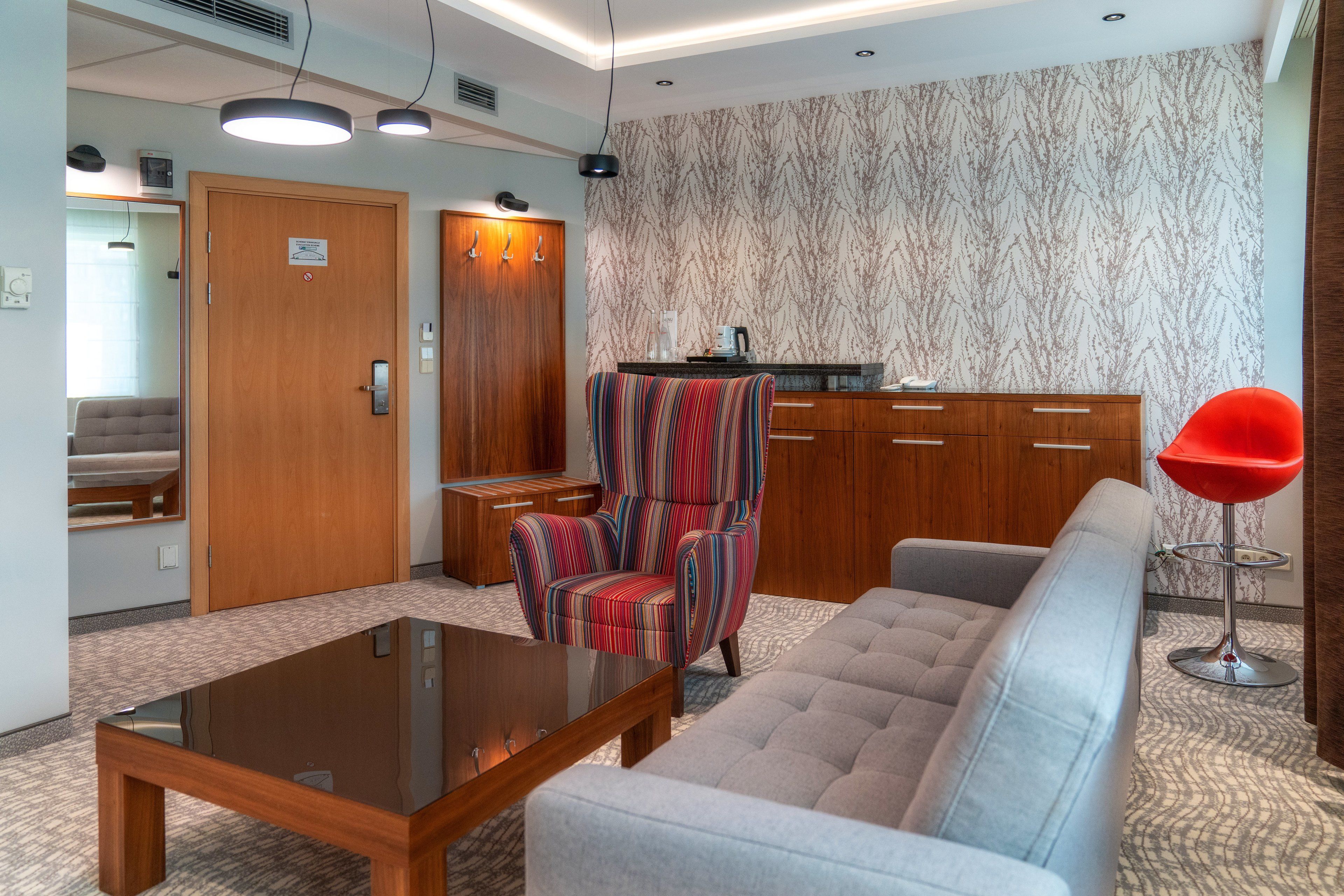 A modern hotel room with a sofa, a striped armchair, a wooden cabinet, and a small kitchenette.