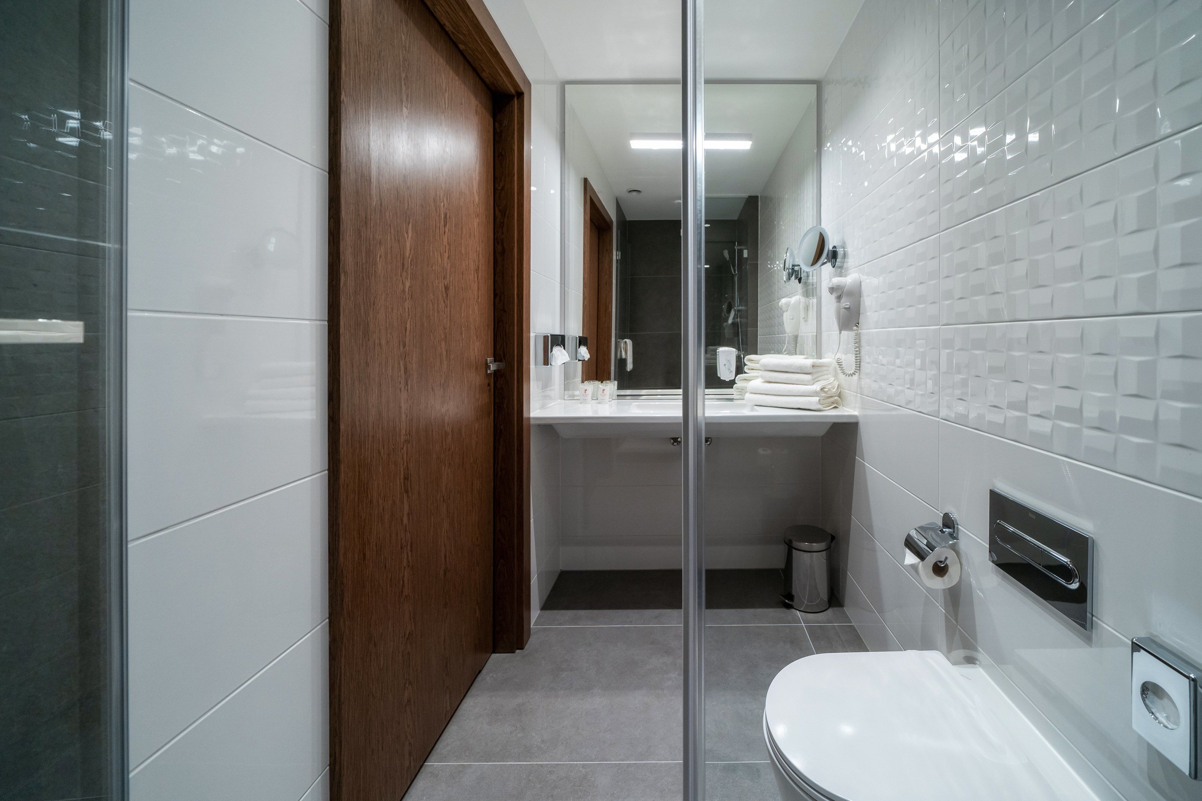 Un baño moderno con mampara de vidrio, puerta de madera, inodoro blanco, lavamanos con toallero y espejo.