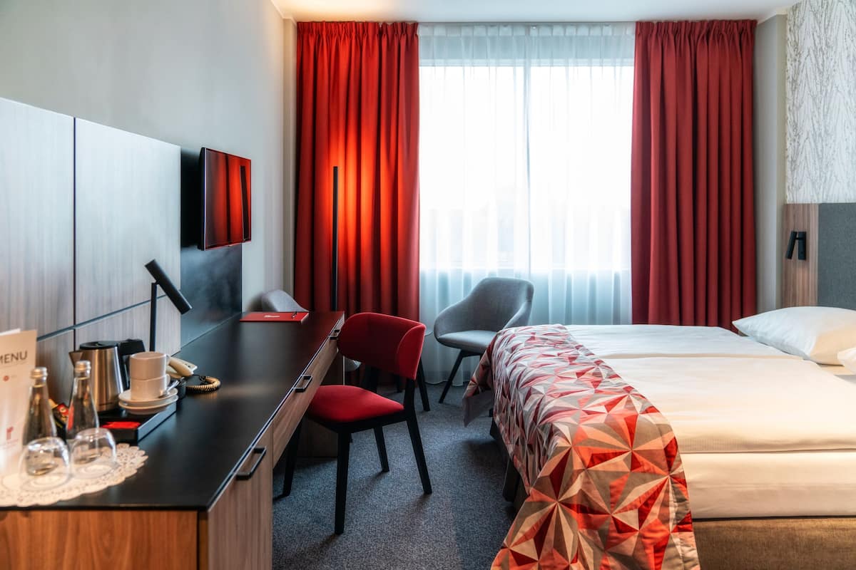 Une chambre d’hôtel avec un lit, un bureau et une chaise, une télévision et une fenêtre avec des rideaux rouges.