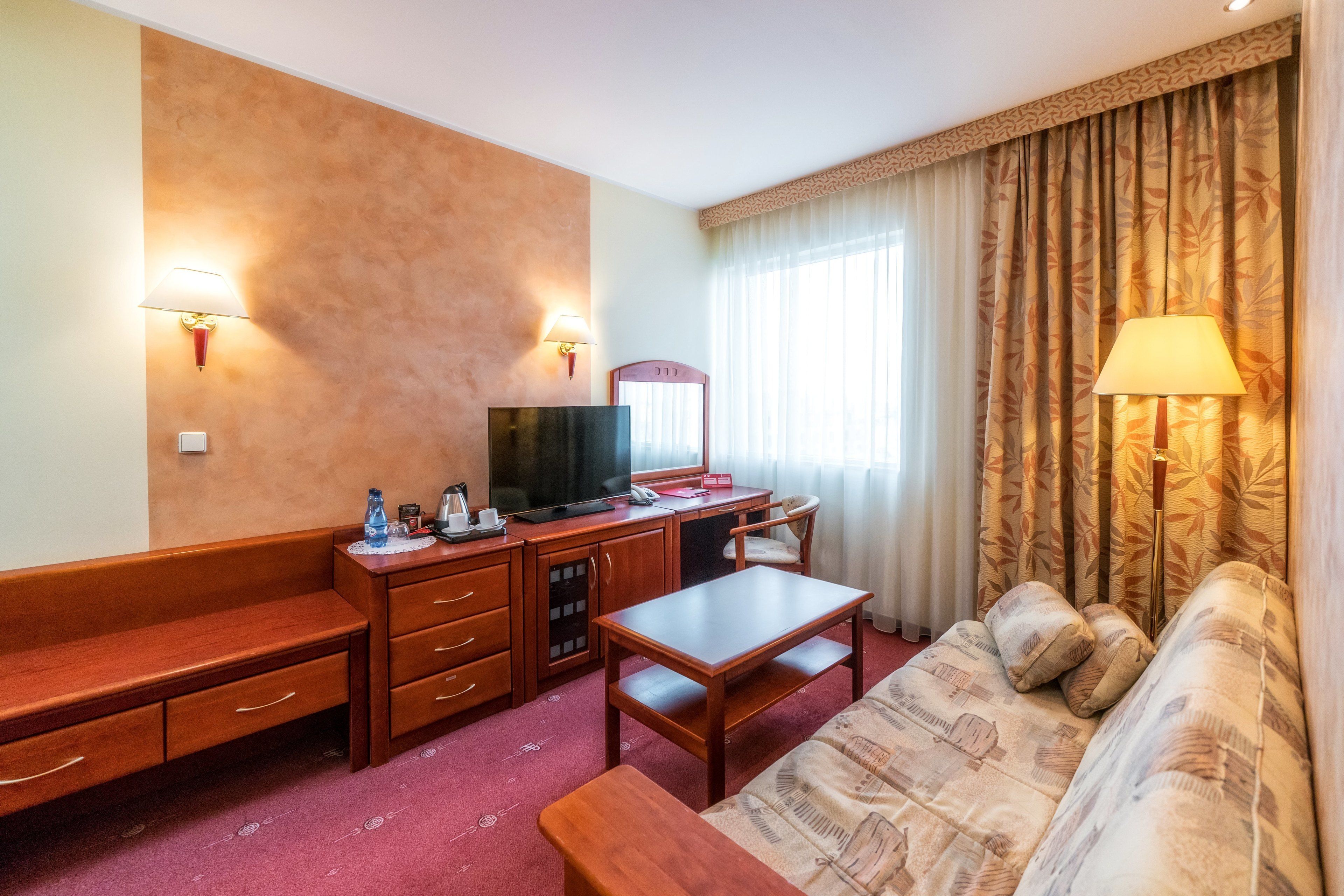 A hotel room with a wooden desk, a television, a sofa, and a patterned couch.