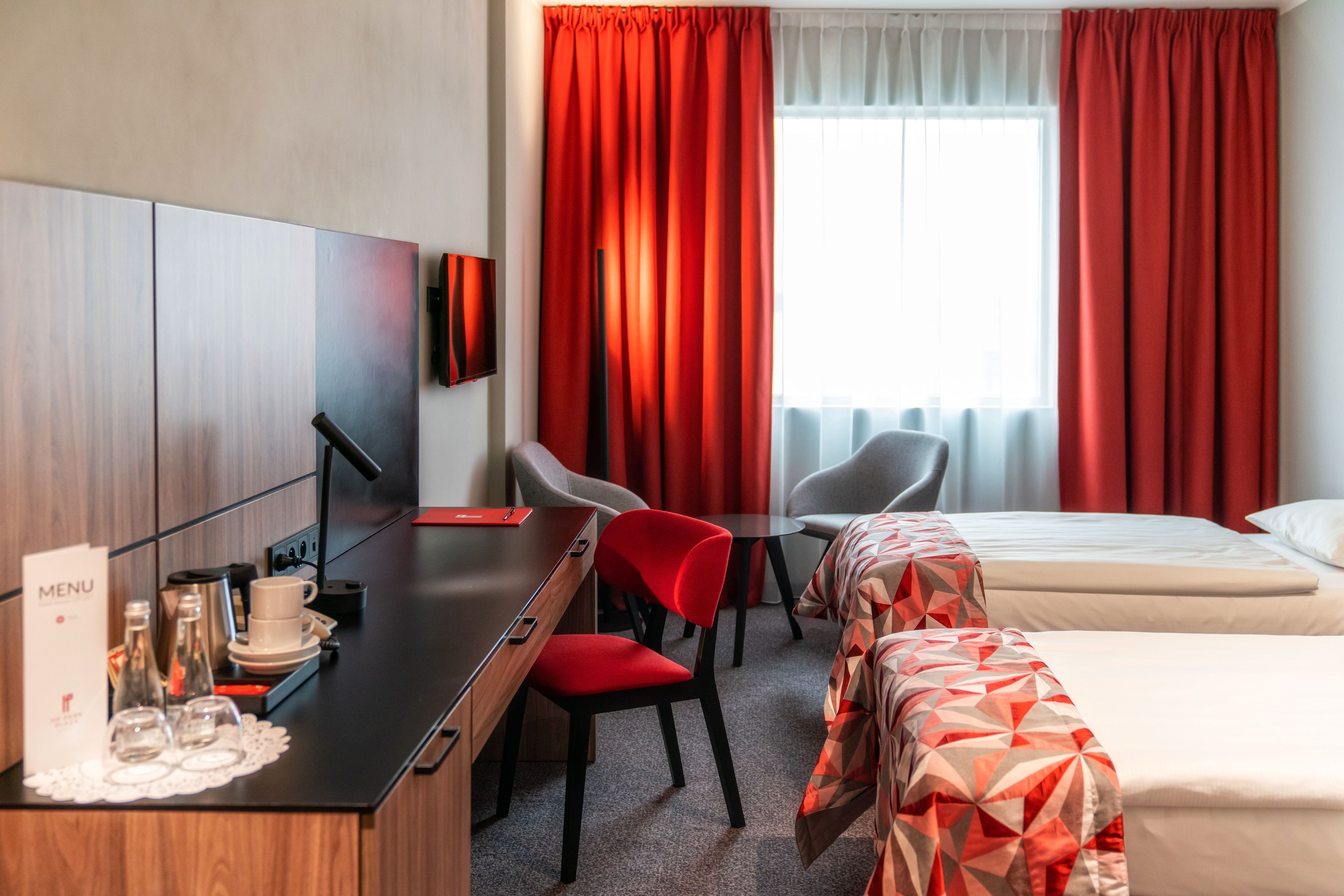 A hotel room with a desk, two chairs, a bed, and a window with red curtains.