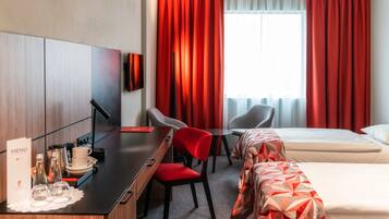 A hotel room with a desk, two chairs, a bed, and a window with red curtains.