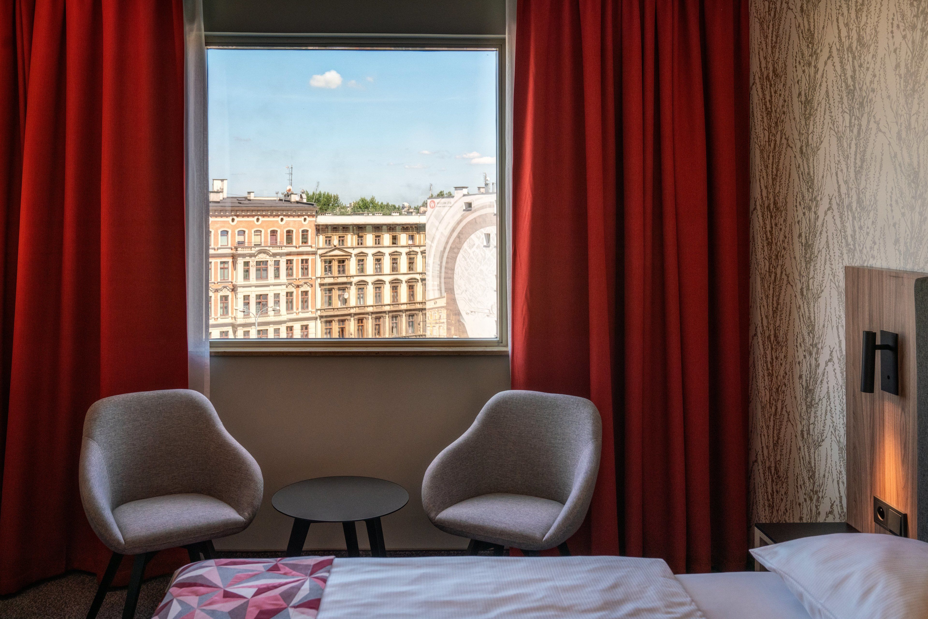 A hotel room with a bed, two armchairs, a small table, and a window with red curtains.