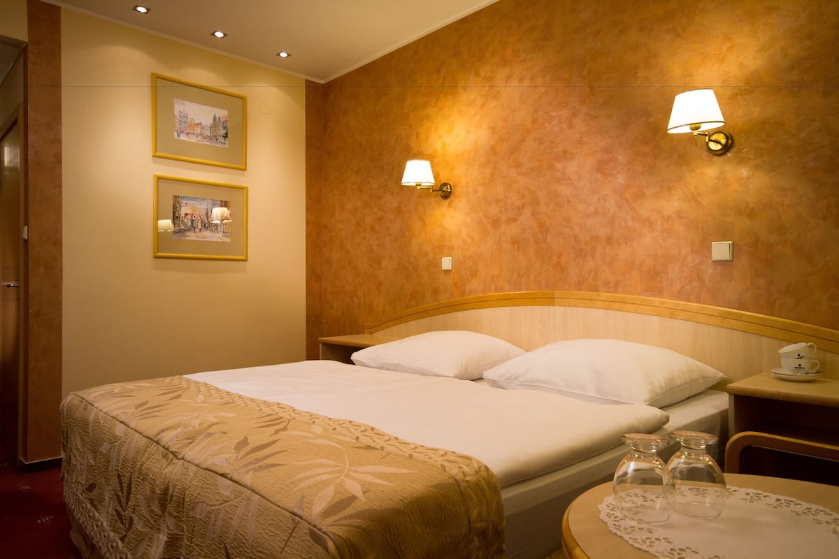 Une chambre d’hôtel comprenant un lit, deux tables de chevet, une petite table avec une tasse et une soucoupe, et deux lampes fixées au mur.