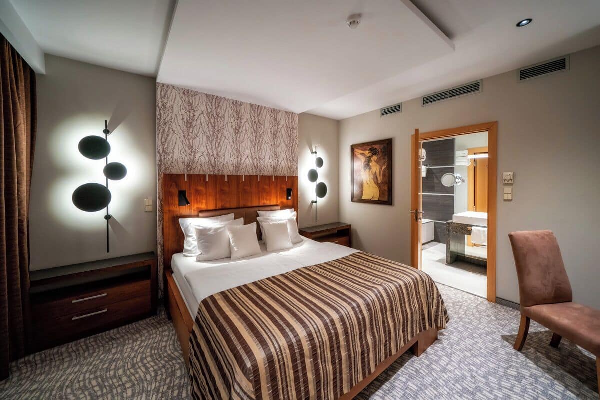 Une chambre d’hôtel avec un grand lit, une tête de lit en bois, un luminaire fixé au mur et une salle de bain visible à travers une porte ouverte.